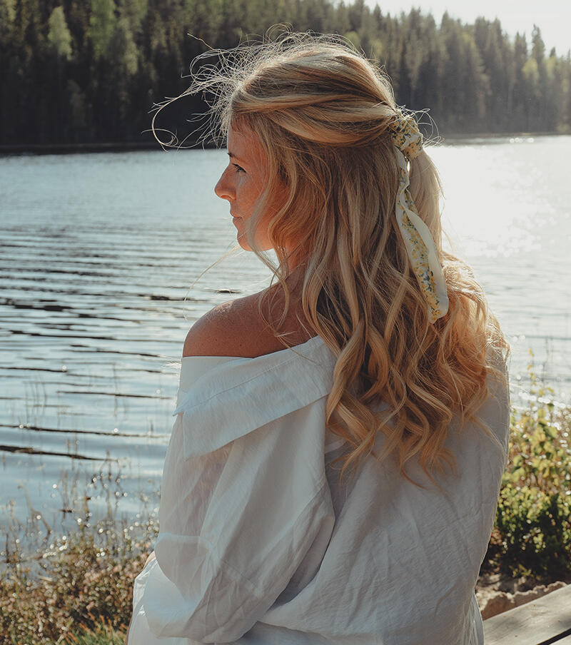 Kvinna med Cendre Ash Blond Mix extensions i en håruppsättning på en bänk framför havet 